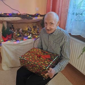 Zdjęcie przedstawia mieszkańca Domu przy szopce bożonarodzeniowej z prezentem mikołajkowym.